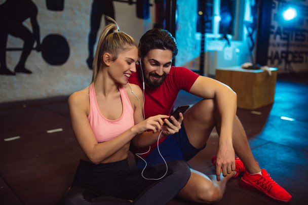 Молодая спортивная пара, пользующаяся мобильным телефоном в спортзале. Друзья слушают музыку в спортзале
. - Фото, изображение