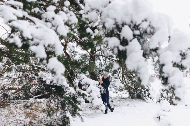 Μια νεαρή, όμορφη κοπέλα ποζάρει κάτω από ένα μεγάλο πεύκο καλυμμένα με χιόνι ΦΟΡΕΣ - Φωτογραφία, εικόνα