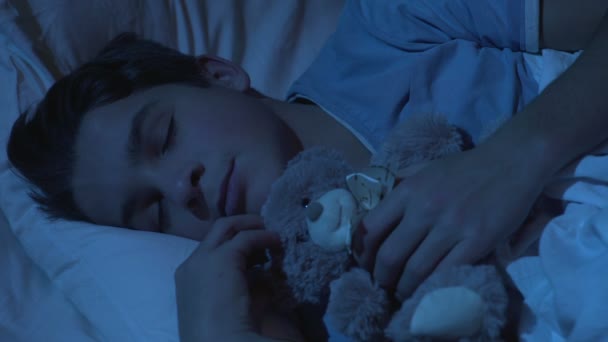 Cute teenage male sleeping in bed with teddy-bear toy, childhood, sweet dreams - Materiał filmowy, wideo