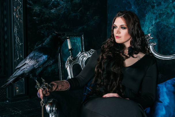 junge schöne kaukasische Frau mit schwarzem Raben auf der Hand sitzt auf dem Sofa - Foto, Bild