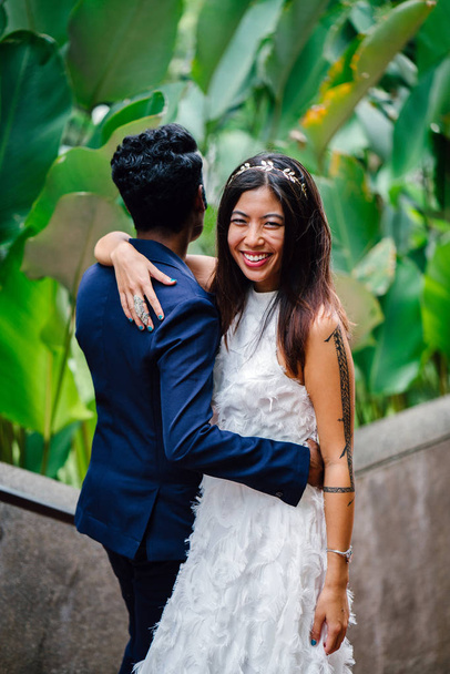 An attractive couple of different races stand together in park in day in Asia. They are taking wedding photographs. One is an Indian man, the other a Chinese woman. - Photo, Image