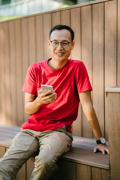middle-aged Asian man talking on his phone in a park. He is sitting on a wooden structure and smiles as he types on his phone at sunset. He is dressed comfortably in a tee shirt and loose pants. - Foto, immagini