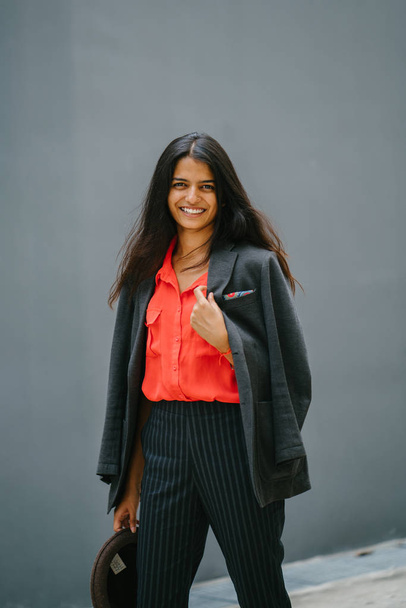 Portrait of a fashionable Indian Asian woman against a plain grey background. She is smiling and wearing a smart orange blouse and black pinstripe pants with a jacket draped over her shoulders. - Foto, Imagen