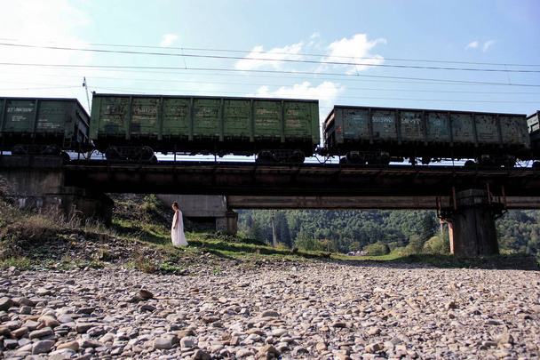 Een meisje in een witte jurk loopt onder een brug. Bewegende trein. Vracht trein met wagons. De trein beweegt tegen de achtergrond van de bergnatuur. Mooie jongedame in witte jurk. - Foto, afbeelding