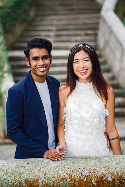  Indian groom holds the wife of his Chinese wife and stand side-by-side in a park. The interracial couple are taking wedding photographs under an arch in the day and are smiling. - Foto, immagini