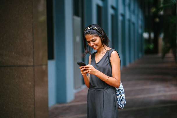 Portrait of young and attractive Indian Asian woman standing in the sun against a dark background in the city. She's standing and smiling in the daytime with her smartphone in hand. - Foto, Imagen