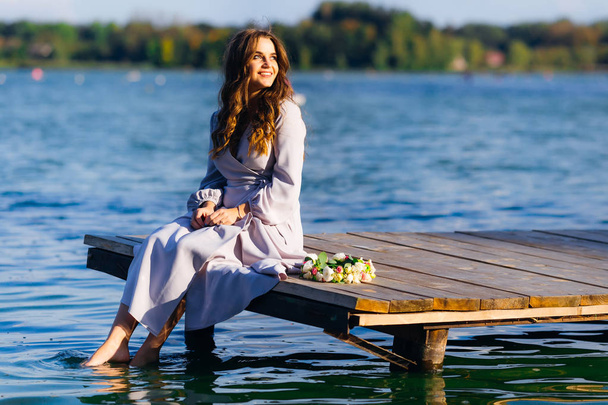 かわいい女の子の木製の桟橋に座っているし、心から水と笑顔で彼女の足を浸す - 写真・画像