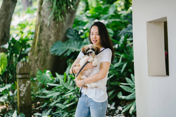 Portrait of a young Pan Asian woman carrying her dog and smiling for the camera in a green park on a warm day in the park. The dog is a toy breed shih tzuh. - Foto, Imagen