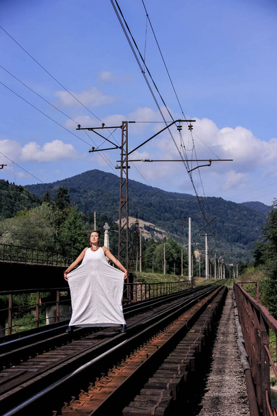 青空をバックに白いドレスの女の子。ジョージア州の緑の山々 の背景に若い女性は。女性は、鉄のレール上に立っています。電車を待っています。ジョージア州の旅. - 写真・画像