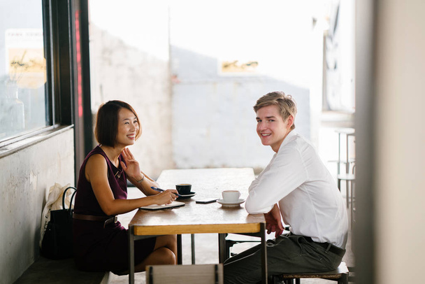 An Asian woman is interviewing a young Caucasian white man for an internship job in Asia. They are sitting at a desk in the shade outside in the day and talking over coffee. - Foto, Bild