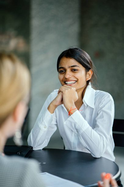  young and attractive Indian Asian woman is interviewing for a job. She is dressed professionally in a white shirt and is sitting and talking to her interviewers. She is confident and relaxed. - Foto, Imagen