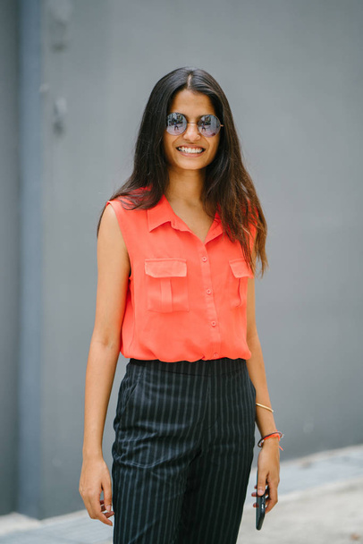 Portrait of a young and fashionable North Indian Desi Asian woman. She has a light complexion and is wearing an orange shirt, black pants . She smiles as she uses her smartphone. - Photo, image