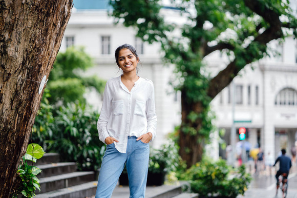 Attractive and young Indian woman standing next to a tree in the city in the day. She's in a casual white blouse and jeans. There is old colonial building and traffic in the background. - Фото, зображення