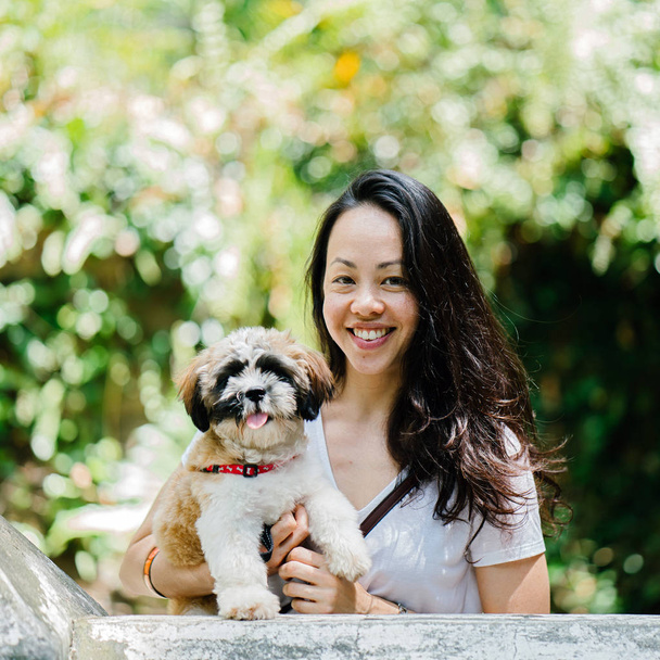 Portrait of a young Pan Asian woman carrying her dog and smiling for the camera in a green park on a warm day in the park. The dog is a toy breed shih tzuh. - Foto, imagen