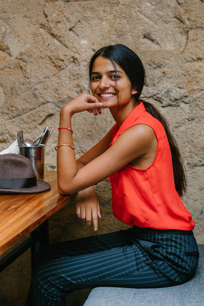Portrait of a young and attractive Indian Asian woman smiling at the camera. She is seated and has a brown fedora cap . She is dressed in smart casual and is relaxed and cheerful. - Foto, Imagem