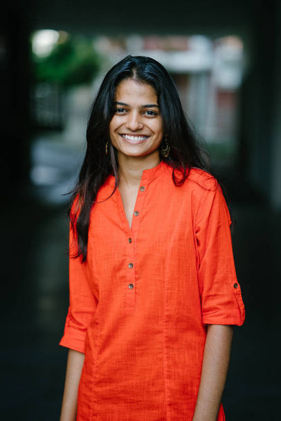 Viehättävä ja nuori intialainen nainen hymyilee kaupungin taustaa vasten. Hänellä on etninen oranssi asu.
. - Valokuva, kuva