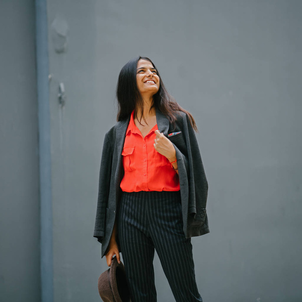 Portrait of a fashionable Indian Asian woman against a plain grey background. She is smiling and wearing a smart orange blouse and black pinstripe pants with a jacket draped over her shoulders. - Фото, зображення
