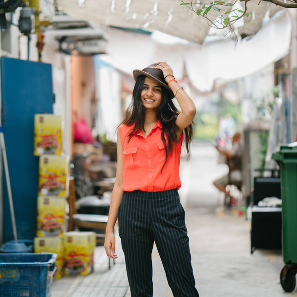 Portrait of a young Indian Asian model wearing a fedora hat standing in an alley in Asia. She is smiling and looking away in the day, and is elegant, pretty and tall. - Φωτογραφία, εικόνα