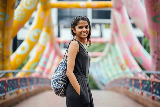 Young, attractive Indian (Asian) woman tourist crossing a colorful bridge along the Singapore river. She is wearing a bag with print. We see her back and she walks across the bridge. - Photo, image