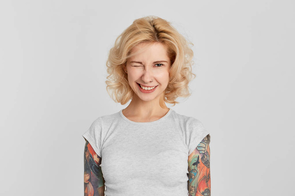 Портрет молодой девушки носит белую футболку с жемчужным носом и татуировкой, улыбается и показывает зубы. Подмигни. Выражение лица. Изолированный над белой стеной
 - Фото, изображение