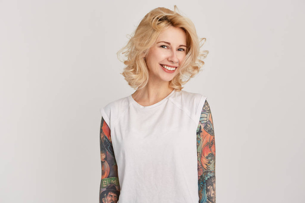 Сором'язлива і усміхнена блондинка з сережкою в носі і татуюваннями над руками дивиться прямо в камеру. Ізольовані над білою стіною
 - Фото, зображення