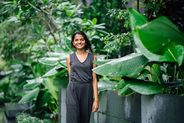 Candid portrait of a young Indian woman standing in a lush green garden. She's dressed in a professional grey smock jumpsuit and smiling. - Foto, Imagen