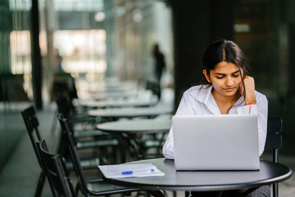 young Indian Asian woman is typing and working in a laptop in the day (applying for a job, etc). She is wearing a professional, crisp white shirt and she is smiling as she types on her laptop. - Foto, Imagen