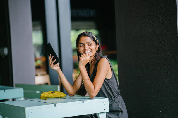 Candid portrait of an attractive and young Indian Asian professional woman reading her tablet or e reader. She is in warm, cosy cafe or coworking space. She is smiling while she reads her tablet. - Photo, image