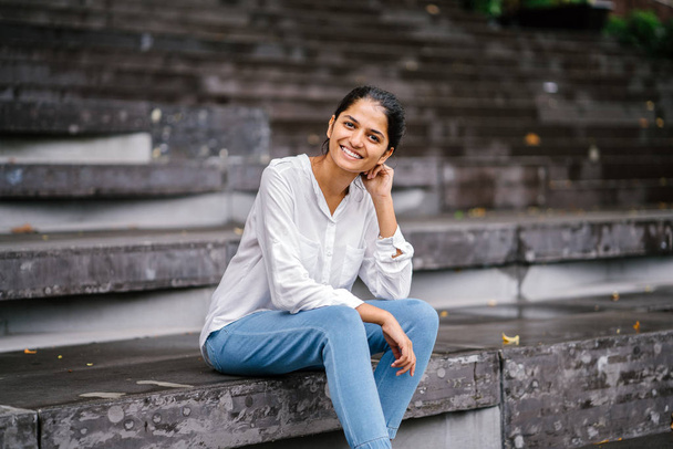 Откровенный портрет улыбающейся привлекательной молодой женщины на серых ступеньках. Индийская азиатка профессиональная, расслабленная и сидящая на ярком солнце
. - Фото, изображение