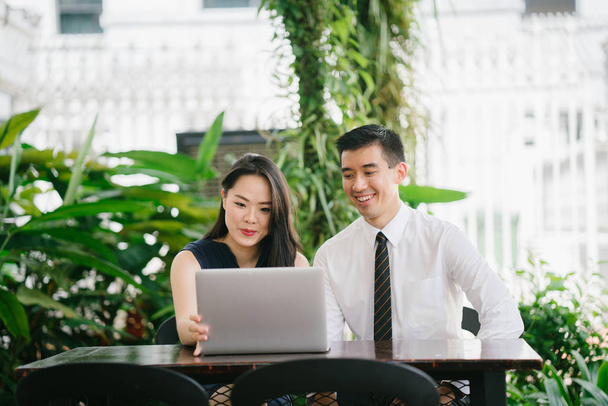 Portrait of two smiling business people (lawyers, consultants, etc) or couple smiling mid discussion over a laptop in an office with greenery in the background. They are focused on the laptop. - 写真・画像