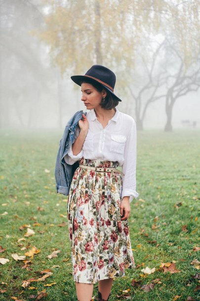 Portrait of young woman wearing white blouse, variegated skirt, denim coat and hat and looking on grass. - Foto, Imagem