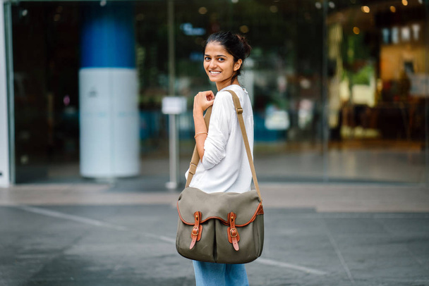 Portrait of a smiling young Asian Indian woman standing and looking over her shoulder in the day in the city. She has a green sling bag over her shoulder. - Zdjęcie, obraz