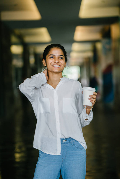 Attractive young Indian Asian woman smiling with a cup of coffee in a campus (dark background). She is dressed in smart casual (entrepreneur in a technology startup) and looking down. - Foto, Imagem