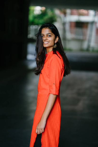 Attractive and young Indian woman smiling against a city background. She's wearing an ethnic orange outfit dress. - Foto, Bild