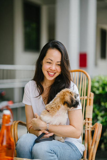  young Pan Asian woman enjoys her weekend with her pet shih tzuh dog. She is sitting in a rattan chair in the sun in a cafe with her dog  - Foto, Imagen