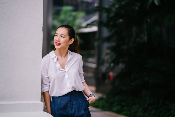 A young Singaporean Chinese woman lounges and relaxes against the wall in the city in Singapore during the day. She is professionally and elegantly dressed in a white shirt and navy pants. - Fotografie, Obrázek