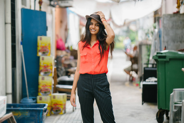 Portrait of a young Indian Asian model wearing a fedora hat standing in an alley in Asia. She is smiling and looking away in the day, and is elegant, pretty and tall. - Fotoğraf, Görsel