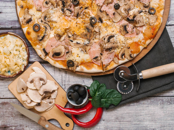 Delicious pizza with tomato sauce, mozzarella, ham, olives and mushrooms served on wooden pizza plate with napkin, pizza cutter, chilli peppers, shinach leaves, knife and sliced mushrooms on chopping board on wooden planks background - Photo, Image
