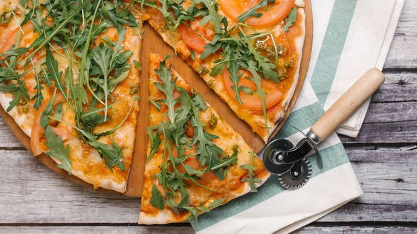 Pizza kesici ve çizgili peçete ile ahşap pizza plaka üzerinde Pizza domates sosu, pesto sos, mozzarella peyniri, roka ve domates ile servis - Fotoğraf, Görsel