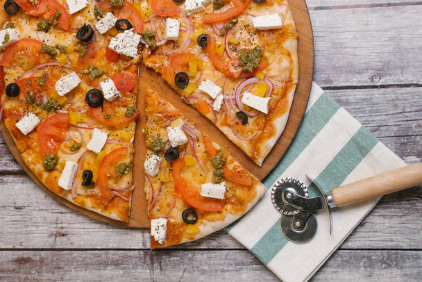 Пицца с томатным соусом, песто соус, моцарелла, помидоры, сладкий лук, оливки, желтый перец и сыр фета подается на деревянной тарелке пиццы с полосатой салфеткой и резак для пиццы
 - Фото, изображение