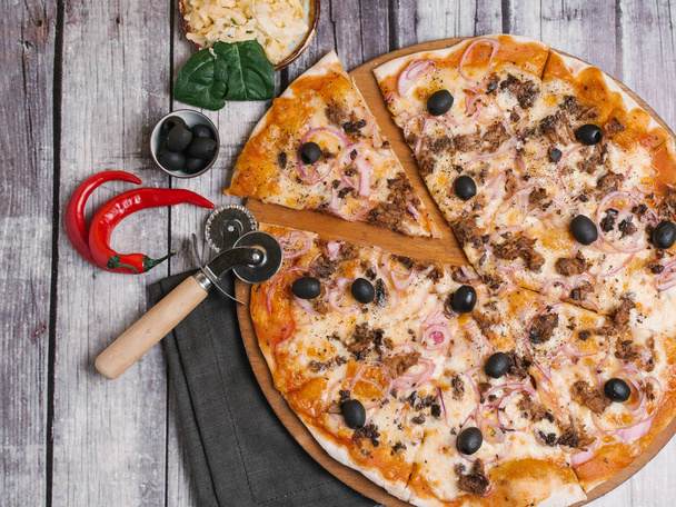 Пицца с томатным соусом, моцарелла, сладкий лук, оливки и фарш подаются на деревянной тарелке пиццы с перцем чили, листья шпината, оливки, салфетки и резак для пиццы на фоне деревянных досок
 - Фото, изображение