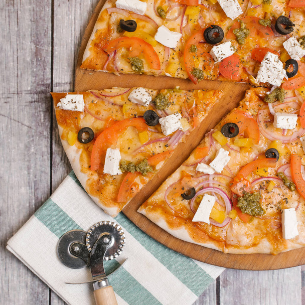 Pizza à la sauce tomate, sauce pesto, mozzarella, tomates, oignons doux, olives, poivrons jaunes et fromage feta servi sur une assiette de pizza en bois avec serviette rayée et coupe-pizza
 - Photo, image