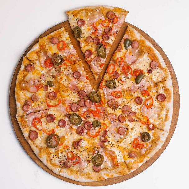 пицца с томатным соусом, моцарелла, ветчина, сосиски, красный перец чили и зеленый перец чили подаются на деревянной тарелке пиццы на белом столе
 - Фото, изображение