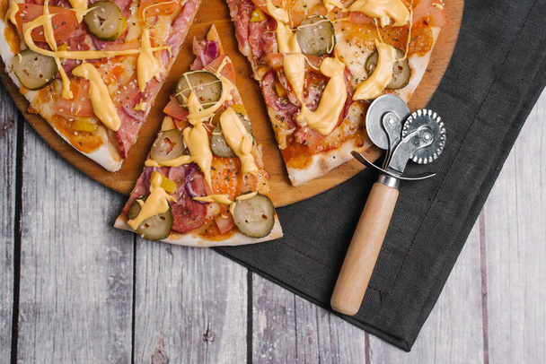 Closeup of pizza with tomato sauce, cheese sauce, mozzarella, tomatoes, sausages, pickles, sweet onions, yellow peppers served on wooden pizza plate with pizza cutter and dark napkin on wooden planks background - Photo, Image