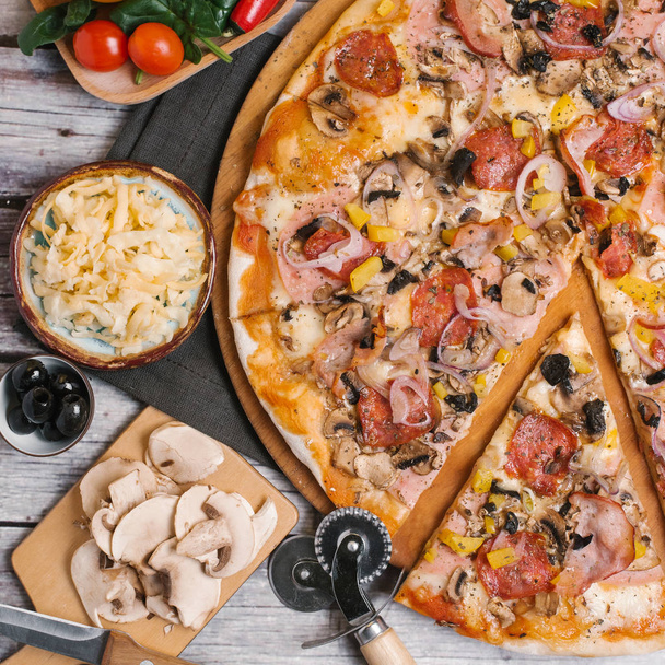 Pizza avec sauce tomate, mozzarella, jambon, saucisses, olives, oignons doux, poivrons jaunes et champignons servis sur une assiette de pizza en bois avec coupe-pizza, serviette et légumes sur des planches en bois fond
 - Photo, image