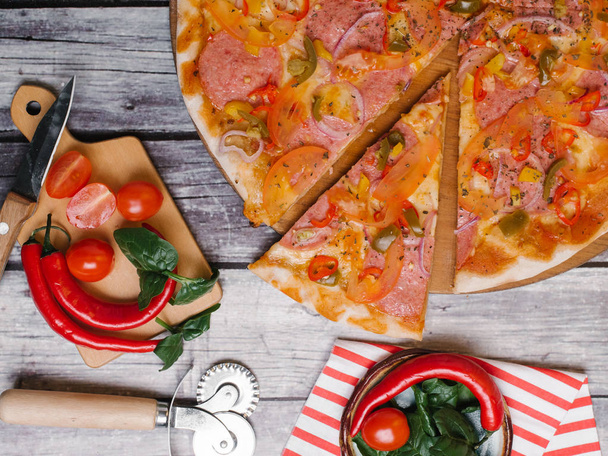 Chilli pizza with pepperoni, green chilli peppers, tomatoes and mozzarella served on wooden plate with pizza cutter, knife and striped napkin on shabby wooden planks background - Photo, Image