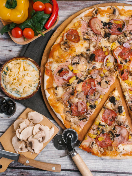 Pizza tomaattikastikkeella, mozzarellalla, kinkulla, makkaroilla, oliiveilla, makealla sipulilla, keltaisella paprikalla ja sienillä, jotka tarjoillaan puisella pizzalautasella, jossa on pizzaleikkuri, lautasliina ja vihannekset puisilla lankuilla tausta
 - Valokuva, kuva