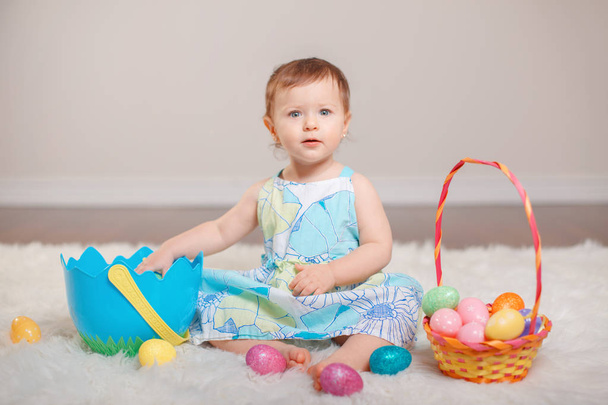 Cute adorable Caucasian baby girl in blue dress sitting on white soft fluffy rug carpet in studio. Kid child holding Easter colorful eggs celebrating traditional holy Christian holiday. - Photo, Image