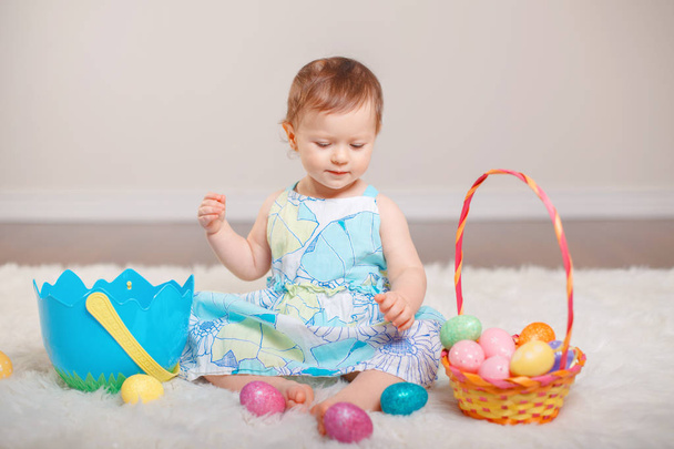 Cute adorable Caucasian baby girl in blue dress sitting on white soft fluffy rug carpet in studio. Kid child playing with Easter colorful eggs celebrating traditional holy Christian holiday. - Photo, Image