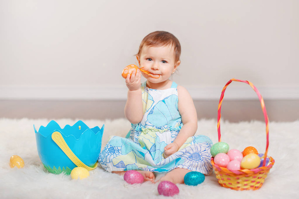 Cute adorable Caucasian baby girl in blue dress sitting on white soft fluffy rug carpet in studio. Kid child playing with Easter colorful eggs celebrating traditional holy Christian holiday. - Photo, Image
